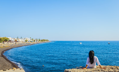Back of woman with black hair sitting on rock, enjoying peacefully the view of mediterranean sea