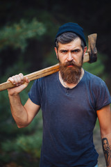 Brutal brunette bearded man in warm hat with a hatchet in the woods on a background of trees