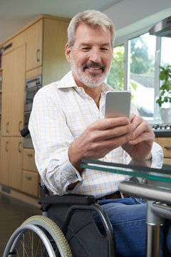 Disabled Man In Wheelchair Texting On Mobile Phone At Home