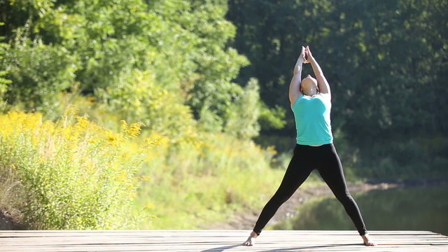 Young beautiful woman in blue tank top practicing outdoors, doing Standing Straddle Forward Bend, Prasarita Padottanasana posture in park on summer day
