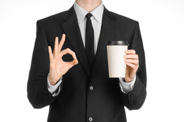 Business lunches coffee theme: businessman in a black suit holding a white blank paper cup of coffee with a brown plastic cap isolated on a white background in the studio, advertising coffee
