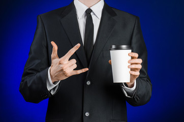 Business lunch and coffee theme: businessman in a black suit holding a white blank paper cup of coffee with a brown plastic cap on a dark blue background isolated in the studio, advertising coffee
