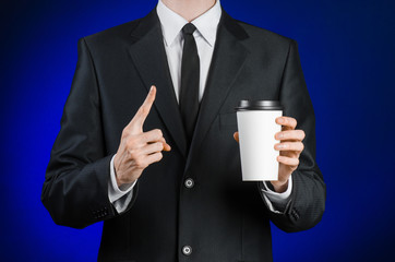 Business lunch and coffee theme: businessman in a black suit holding a white blank paper cup of coffee with a brown plastic cap on a dark blue background isolated in the studio, advertising coffee