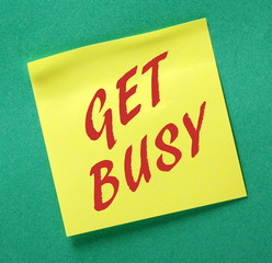 The phrase Get Busy in red text on a yellow sticky note posted on a green notice board as a reminder