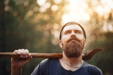 bearded brutal man in warm hat lumberjack with an ax in a forest at sunset, and the thick forest in...