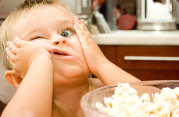 Funny baby bow with .skinned elbow and one wide opened eye on a kitchen background near the bowl of popcorn is making faces - 94096249