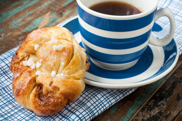 Swedish cinnamon berry bun served on a rustic wooden table with a cup of freshly brewed coffee in a blue and white striped cup and saucer. Coffee and a bun is a tradition in Sweden known as fika.