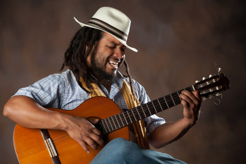 A black Rastafarian man holding a classical guitar with American flag design, wearing a Panama hat, strumming the strings.
