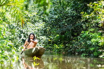 Indigenous adult man skillfully navigating the murky waters of the Ecuadorian Amazon in a traditional wooden canoe carved from a single tree through the dense primary jungle