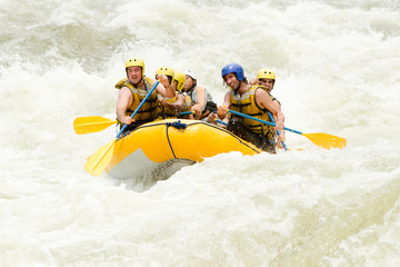 A group of tourists in helmets and life jackets navigate through the white water rapids of a river in Ecuador on an extreme rafting adventure with a guide.