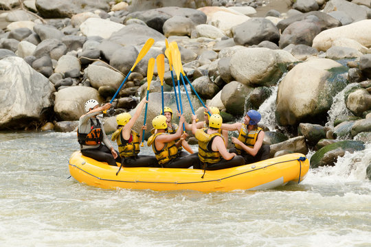 A group of friends in a bright yellow raft paddle through turbulent whitewater rapids surrounded by large gray boulders in Ecuador, wearing life jackets and helmets in an energetic team adventure.