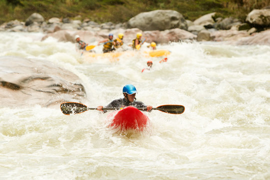 A kayaker in a blue helmet and red kayak paddles through turbulent whitewater rapids viewed from behind in Ecuador, a group of kayakers upstream, capturing the thrill of whitewater kayaking.