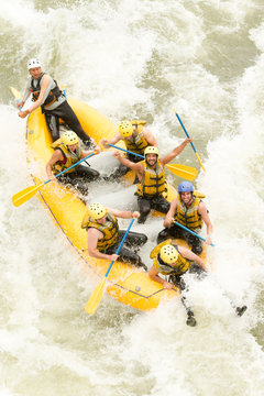 A group of six adventurers in a bright yellow raft paddle through turbulent whitewater rapids, wearing life vests and helmets, splashing through the river with focused teamwork in Ecuador.