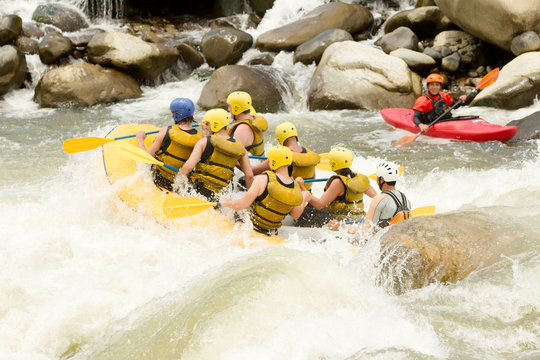 A group in life vests and helmets paddle a yellow raft through turbulent whitewater rapids beside a red kayak on the Pastaza River in Ecuador, through the rushing current.