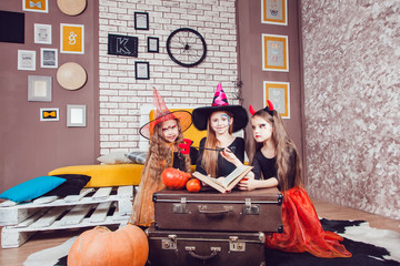 There are three little girls, who are dressed in costumes for Halloween. They are looking like three witches. They are siting near the table with opened magic book.