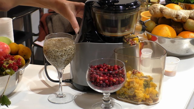 Woman puts some cranberries into working modern electric juice extractor with fresh fruits, seeds and berries at the side