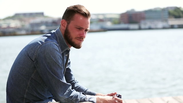 A man in his 20s sitting down wearing a jeans shirt with the sea and city behind him looking down on a sunny summer day.