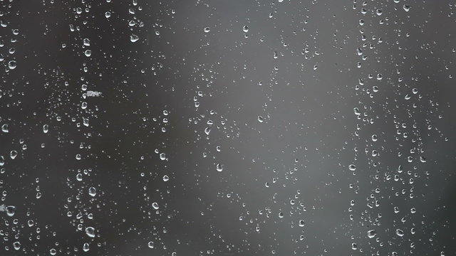 Water drops from melted snow on window glass against grey background. Snowing, stormy weather, movement of snowflakes visible through glass.