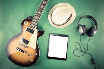 Electric guitar with a tablet, hat and headphones on a green background. Filtered retro image