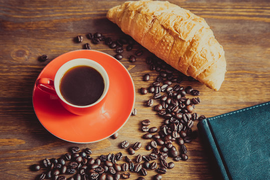Coffee cup and beans, with croissant, notepad  on wooden background, vintage toned