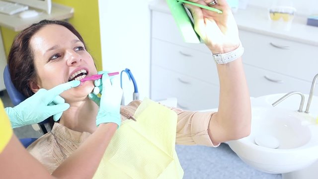 Female dentist showing to her sitting and looking at a mirror female patient how to use dental interproximal brush correctly / camera moving left / close-up