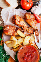 baked chicken legs in garlic marinade with baked potatoes , sauce , tomatoes , bread and herbs on wooden background