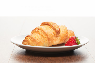 Fresh french croissant and strawberry on white ceramic plate on bright light brown wooden table background