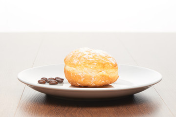 Fresh doughnut with icing sugar and coffee beans on white ceramic plate on bright light brown wooden table background