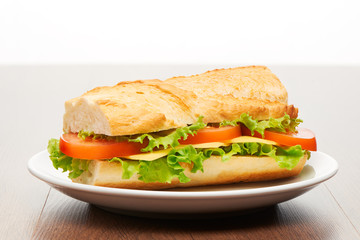 Tomato, cheese and salad sandwich from fresh baguette on white ceramic plate on bright light brown wooden table background
