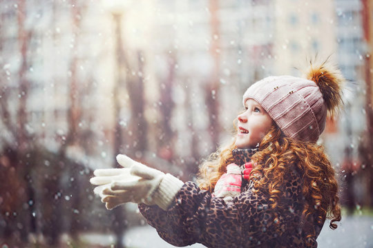 Cute little girl stretches her hand to catch falling snowflakes.