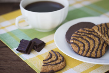horizontal image of a cup of coffee and a plate of oatmeal cookies drizzled with chocolate sitting on a yellow checkered place mat.