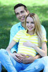 Young pregnant woman with husband sitting on green grass in park