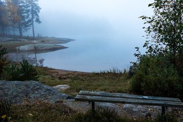 old bench on the shore by a lake covered in fog an early autumn morning
