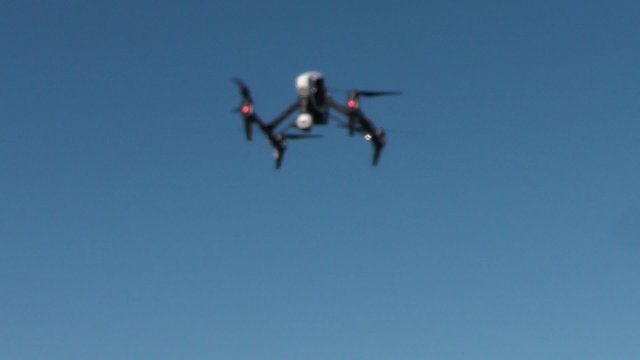 Small drone fitted with camera rises into frame, hovers with camera facing the viewer and then rises to fly out of frame. Shot against a clear blue sky.