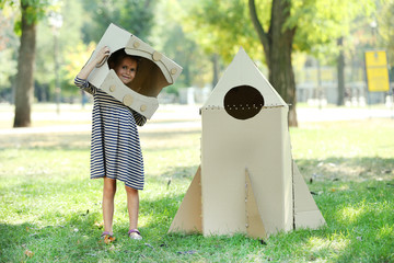 Pretty little girl in box helmet standing near carton rocket in  the park