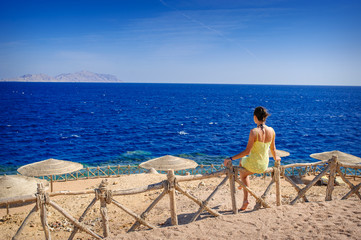 woman in a yellow dress sitting on a fence on the beach and looking at sea