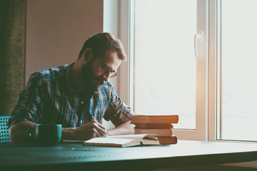 Bearded man writing with pen and reading books at table - 94372682