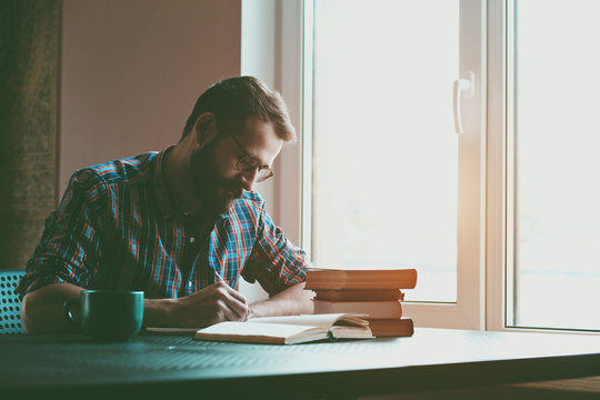 Bearded Man Writing With Pen And Reading Books At Table