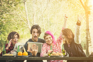 Smiling friends in the park, with food on the table, watching sport game on a tablet pc on a sunny day with their dog - Caucasian people - people, nature, animal, technology and lifestyle concept 