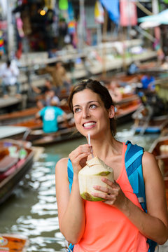 Happy female tourist visiting Damnoen Saduak floating market at Bangkok, Thailand. Woman on Asian travel drinking fresh coconut water.