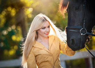 Fashionable lady with yellow coat near black horse in forest. Beautiful young blonde woman with sun rays in hair posing with a friendly horse. Attractive elegant female with horse, sunny summer day