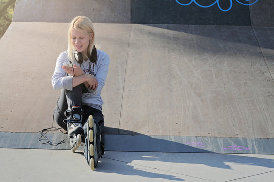 Happy student girl reading text message on smart phone siting on ramp in skate park. Skating and roller sports. Fitness girl taking a break and choosing music for rollerskating