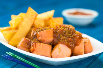 German fast food called Currywurst served with French fries on a disposable plate with party forks in the front (Selective Focus, Focus on the front of the sausage on top)