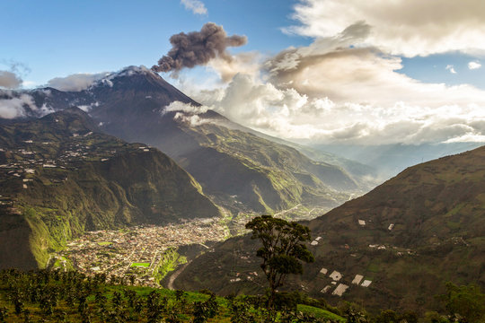 A panoramic view of the Tungurahua volcano releasing an ash plume above the green valley and town of Banos de Agua Santa in the Andes of Ecuador under a dramatic cloudy sky.