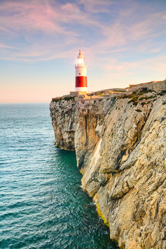 The red-and-white Trinity Lighthouse stands atop the rugged cliffs of Gibraltar overlooking the deep blue sea at sunset, the sky glowing soft pink and orange, a picturesque coastal scene.