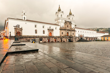 Fototapeta premium The white twin-towered San Francisco Church and its wet stone plaza on an overcast rainy day in the historic colonial center of Quito, Ecuador, reflections on the pavement evoking a quiet serene mood.