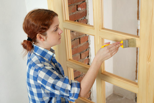 Young female worker painting new wooden door with paintbrush
