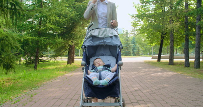 Confident young businesswoman talking on cell phone while walking through a park with her baby boy napping in pram