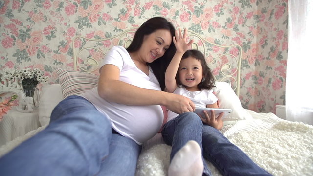 young pregnant Asian woman with her little daughter have fun playing with tablet on the bed