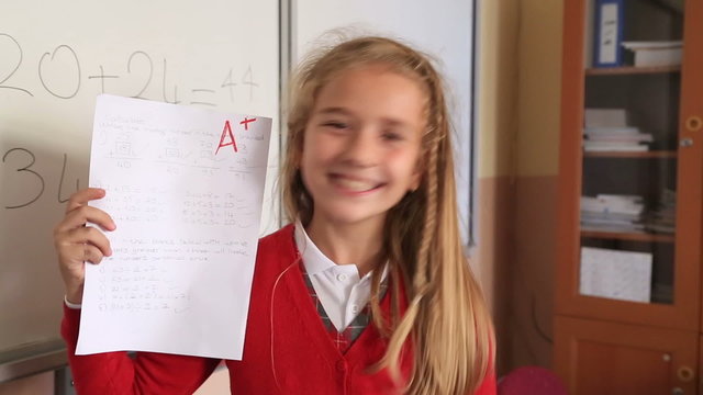 Happy cute little girl showing exam paper with a plus in the classroom